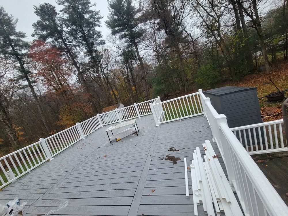 A gray wooden deck with white railing, surrounded by trees with fall foliage; overcast sky.