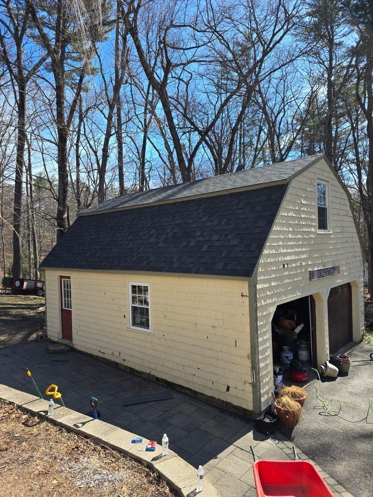 Tan garage with black roof, two garage doors, red door, and windows. Asphalt driveway, trees in background.