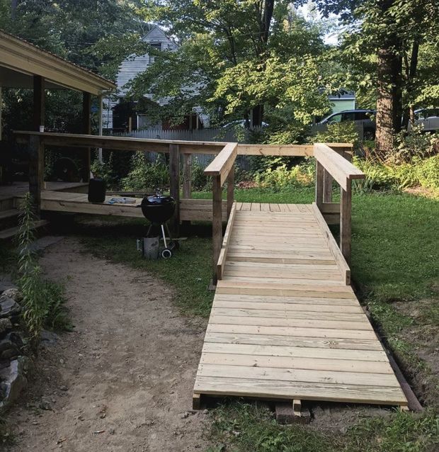 Wooden ramp and deck leading to a backyard, with a grill and trees.