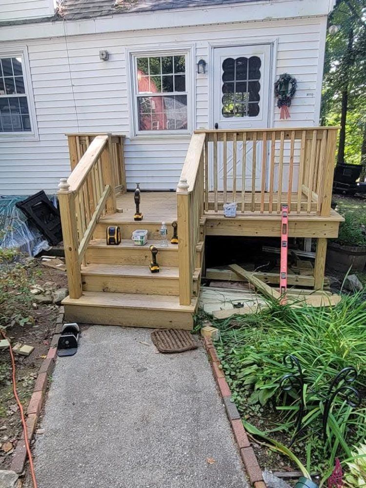 Wooden deck with steps leading to a white house. Handrails and tools visible. Concrete walkway. Green plants surround.