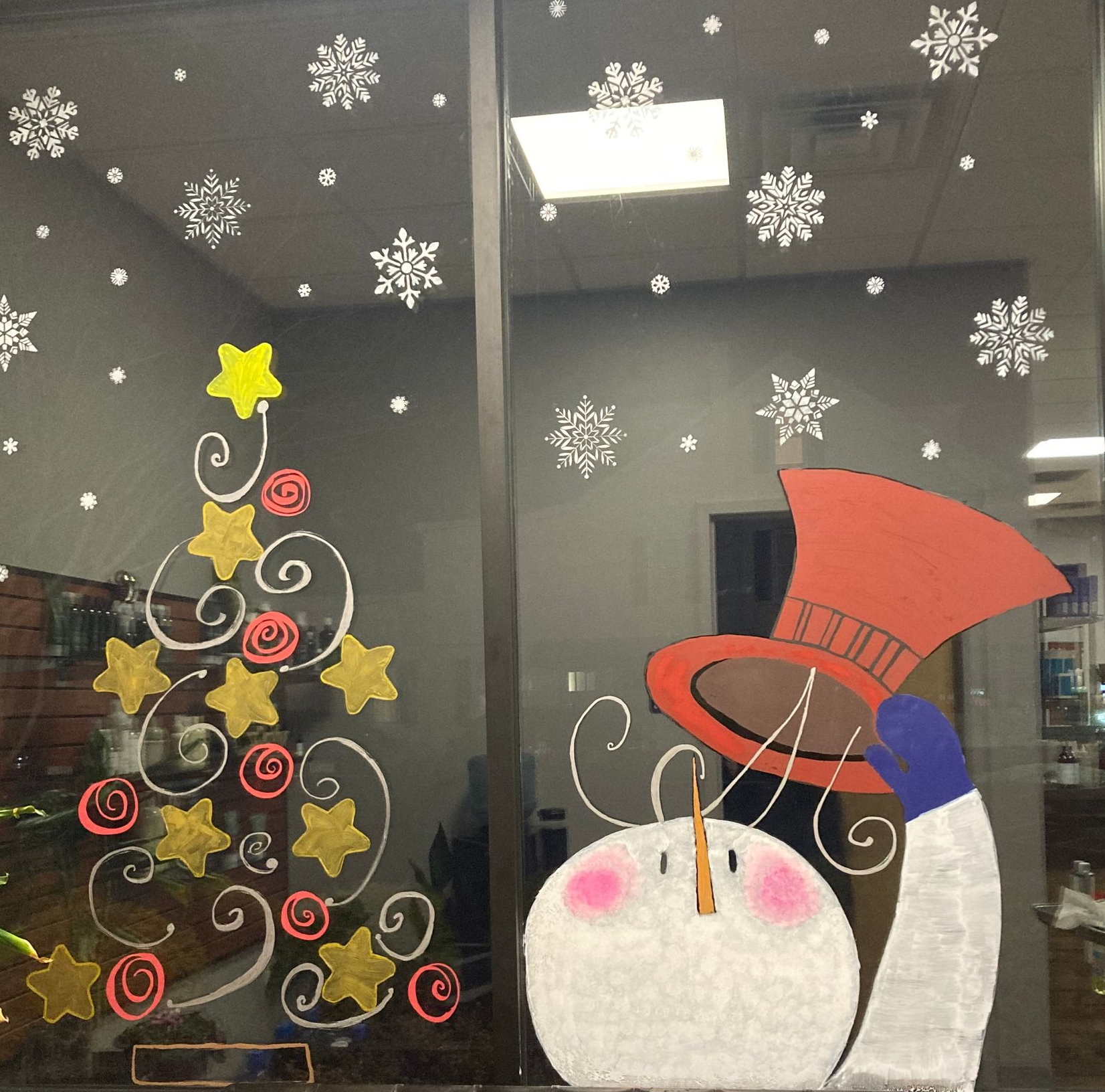 Window display with a painted Christmas tree, snowman, and snowflakes. A brown tufted bench sits below.