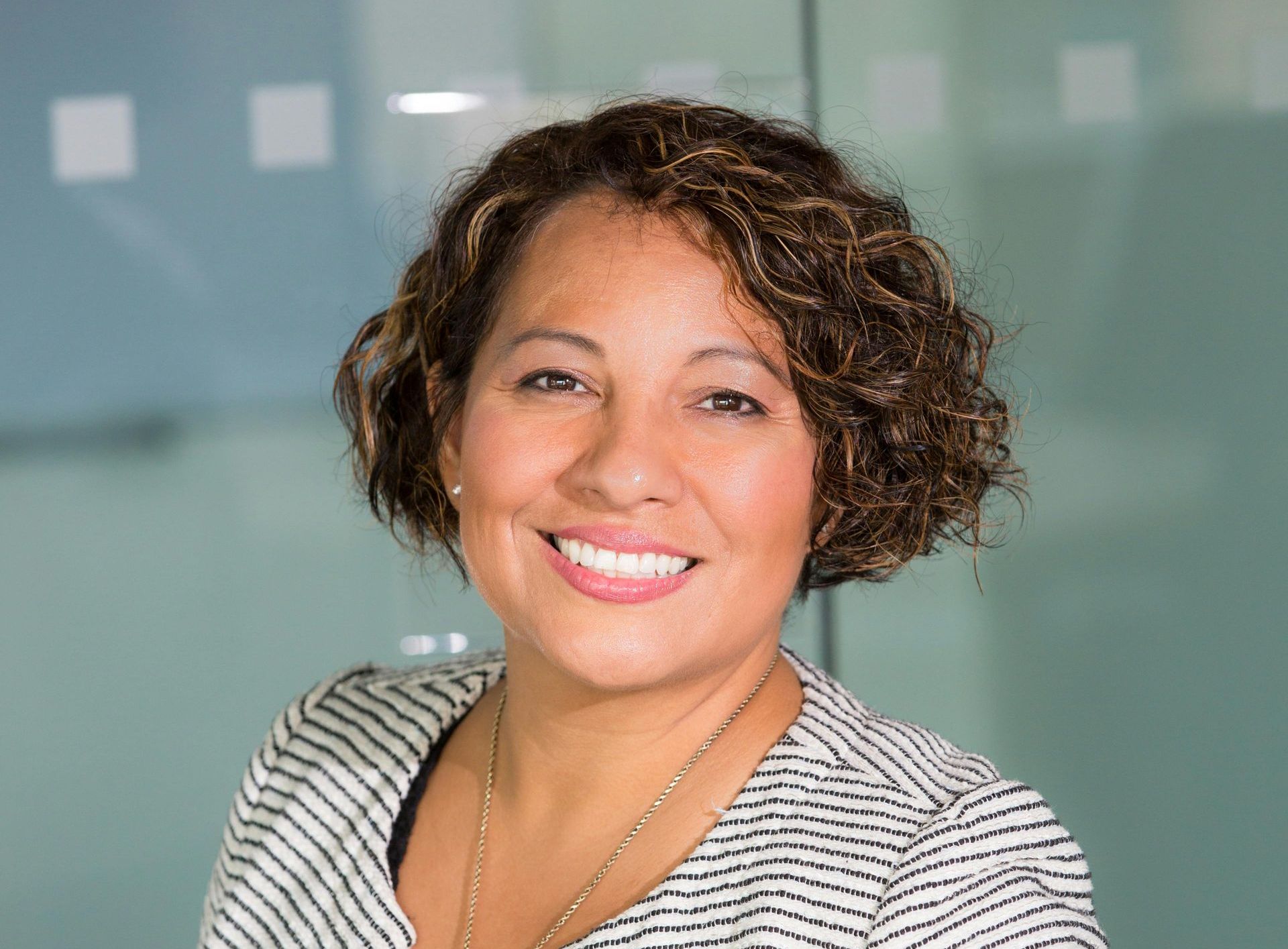 Woman with curly brown hair smiles, wearing striped blazer, in a modern office.