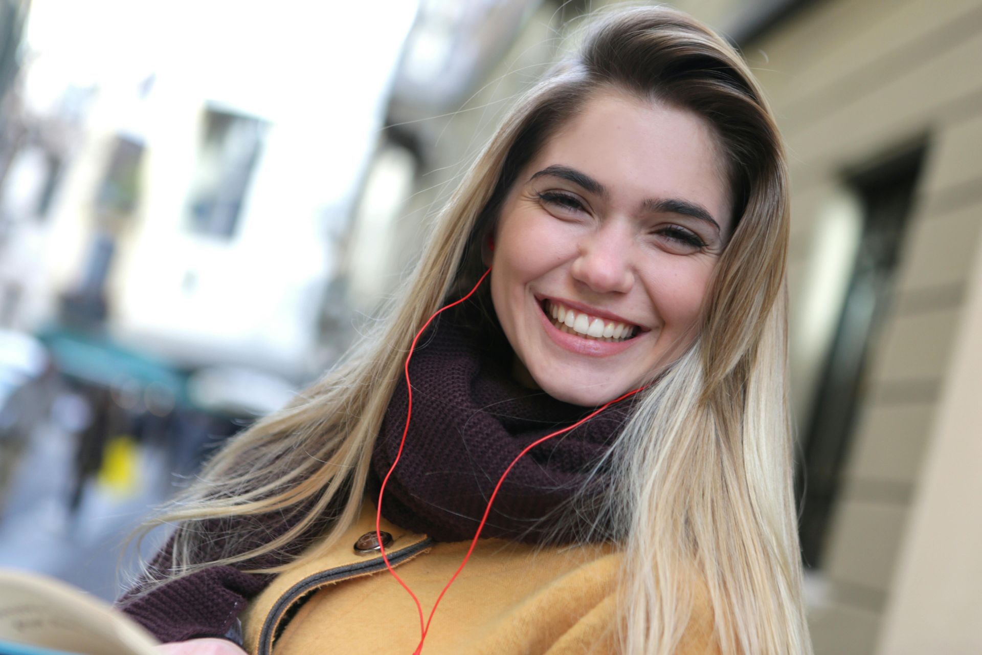 Woman with blonde hair smiles widely, wearing scarf and coat. Red earbuds visible. Outdoors.
