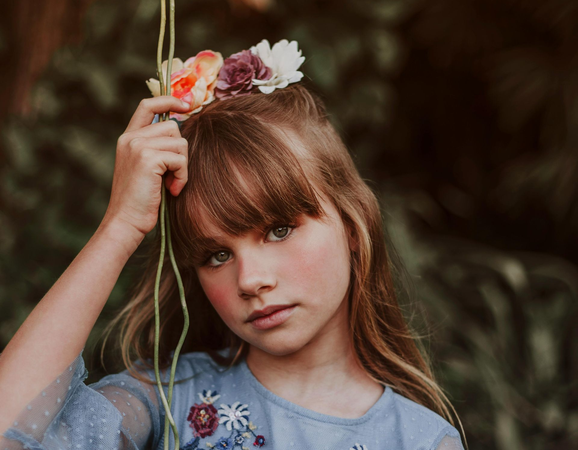 Girl with flower crown and blue embroidered top looking at the camera, holding a string.