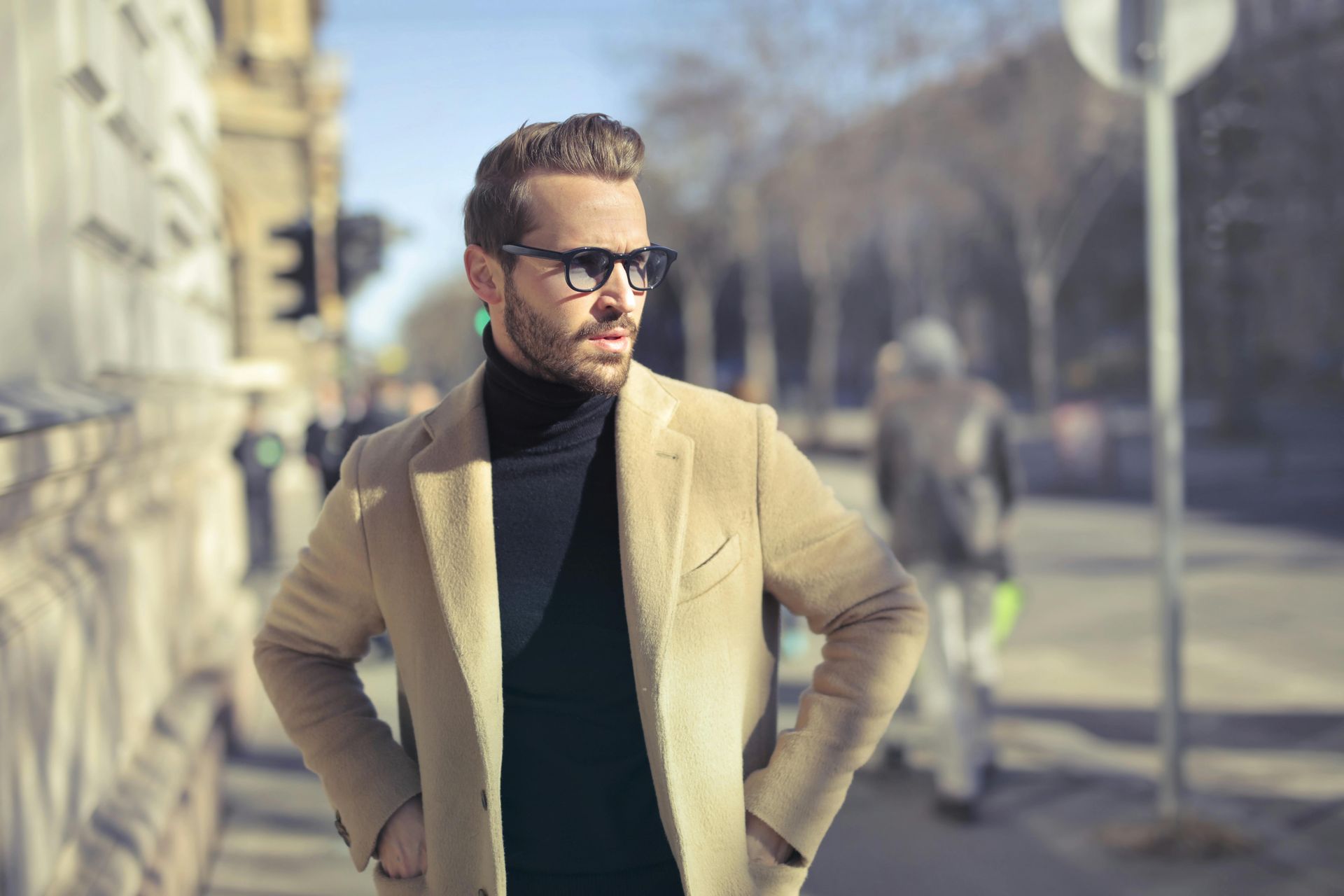 Man in a tan overcoat and sunglasses stands on a city street, hands in pockets, looking off to the side.