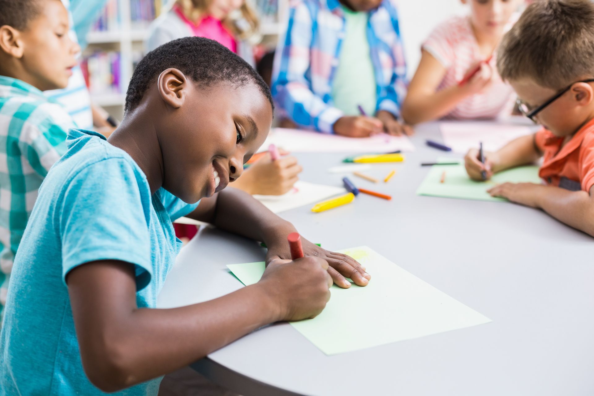 Children at a round table, coloring on paper. Boy in a blue shirt smiles as he colors.