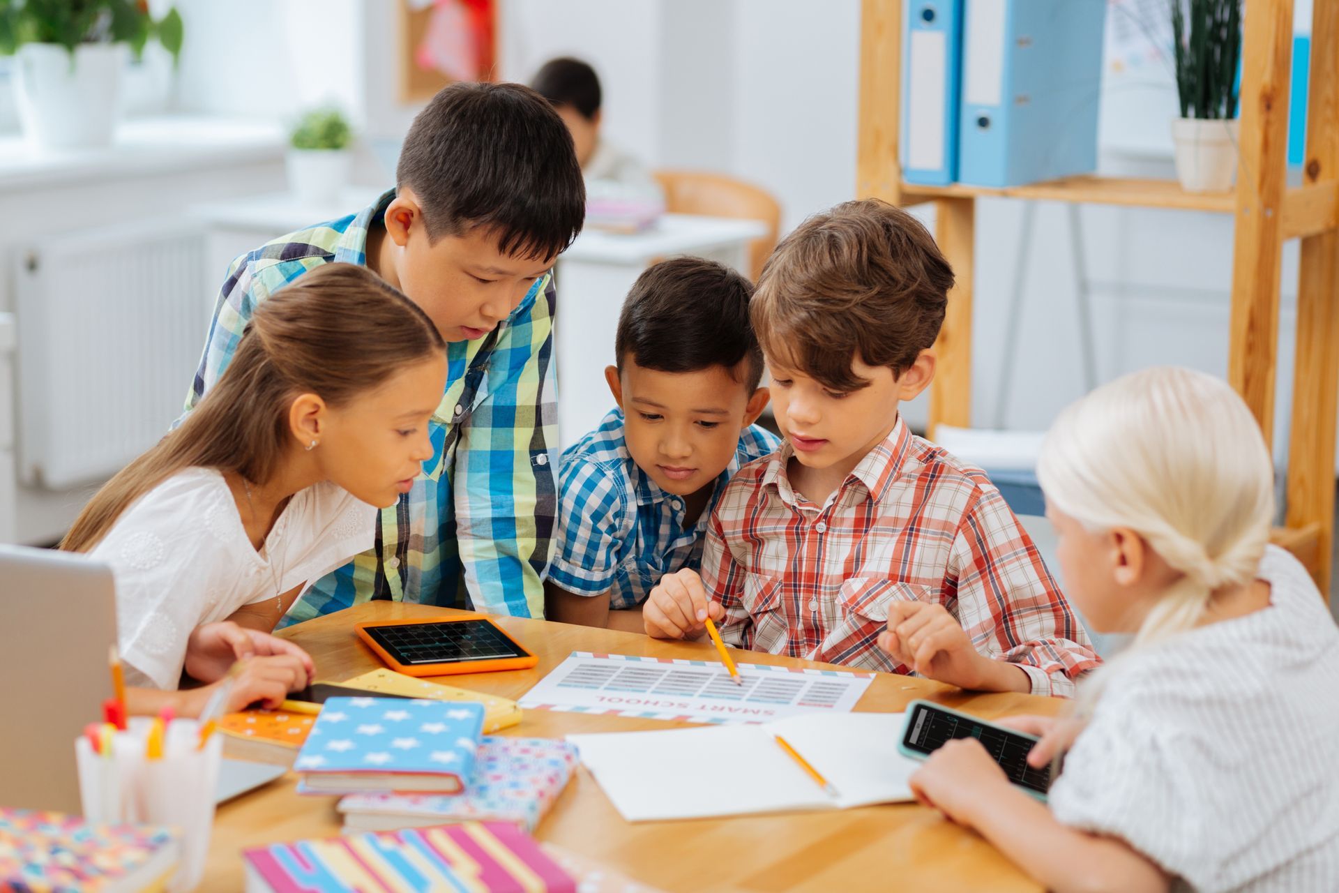 Children huddled around a table, looking at paper. Some point with pencils. Books and a tablet are nearby.