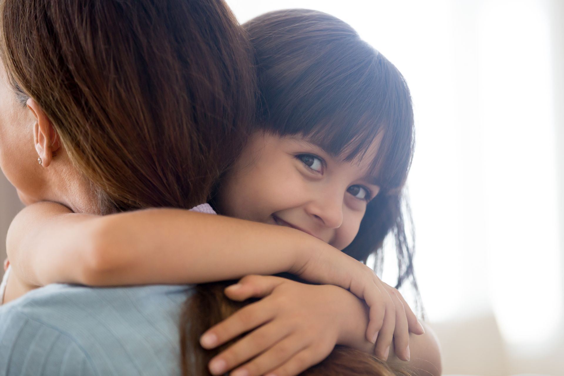 Child hugging a person from behind, smiling. Brown hair, light background.