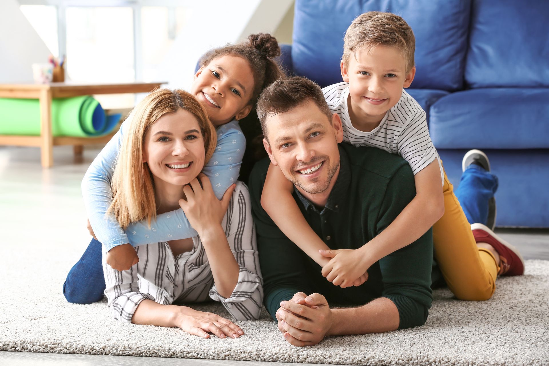 Family of four smiling on a carpet, hugging. Blue couch in the background.