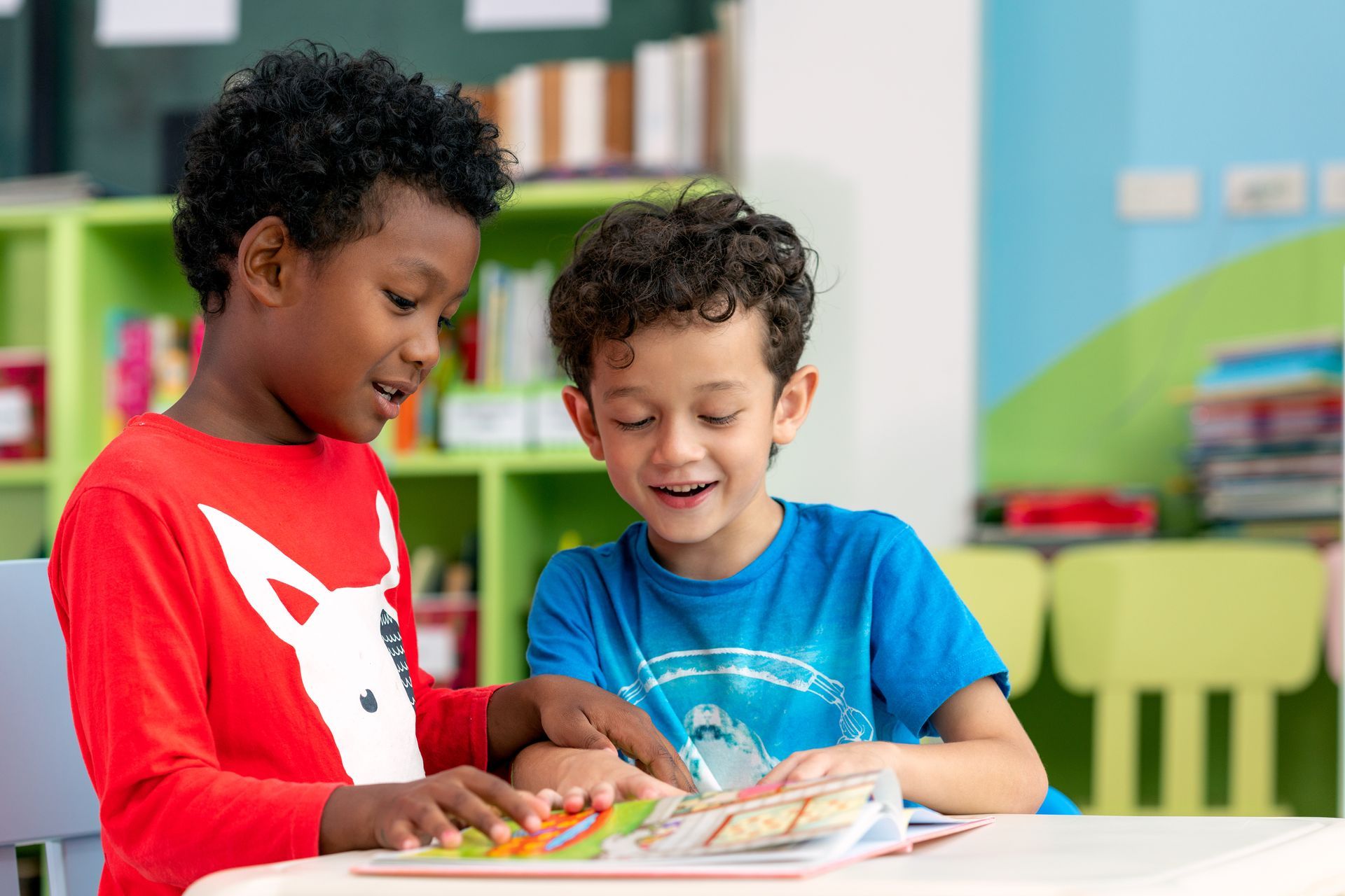 Two young boys reading a book together at a table in a classroom.
