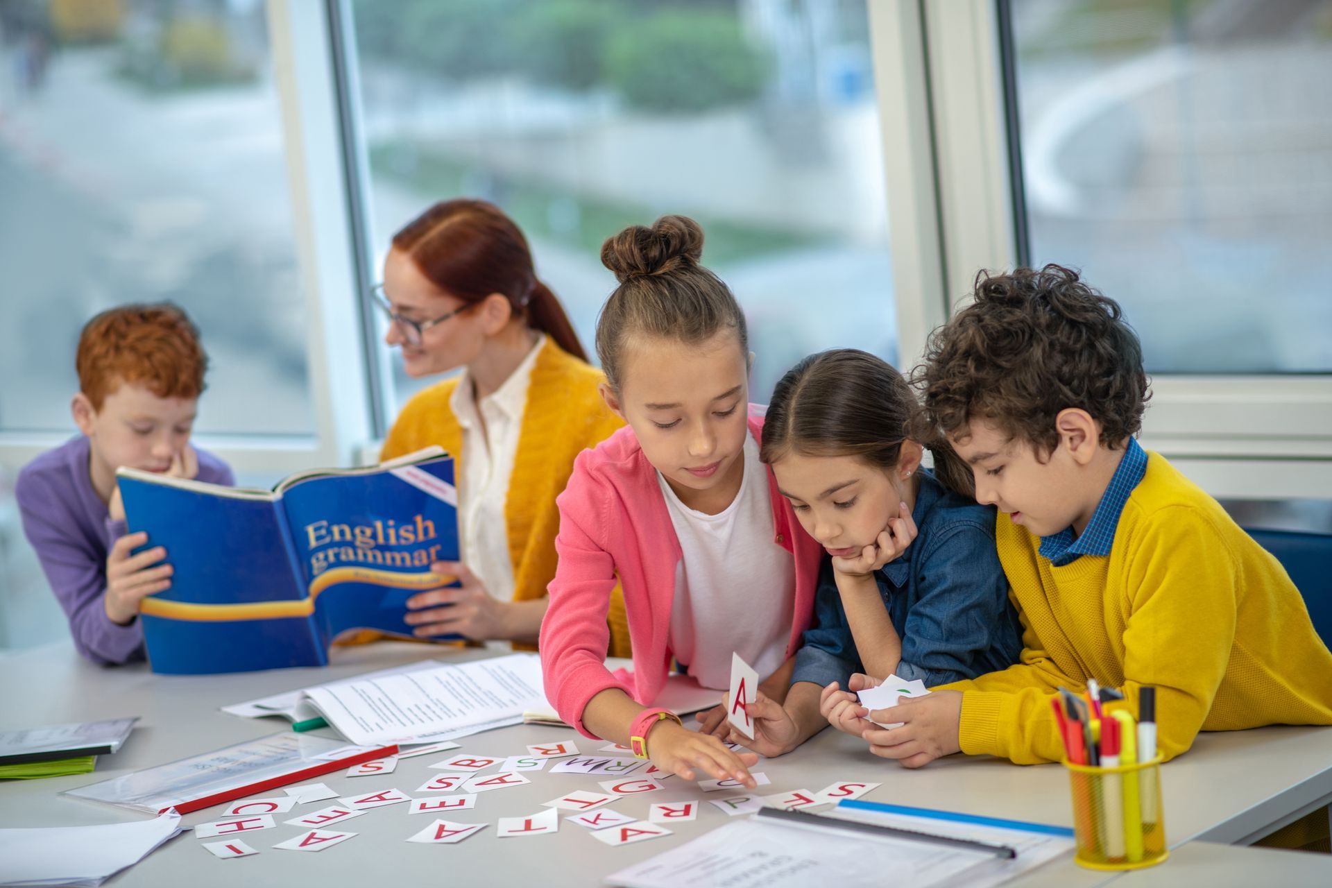 Children and teacher at a table. Students play with cards, teacher reads a book. Class setting.
