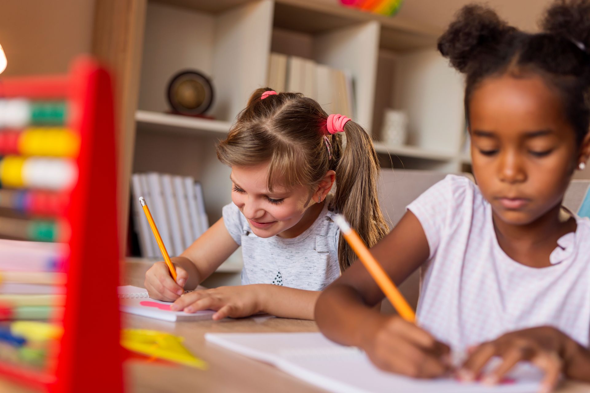 Two children writing at a table, smiling. One has pigtails, the other has buns. Red abacus to the left.