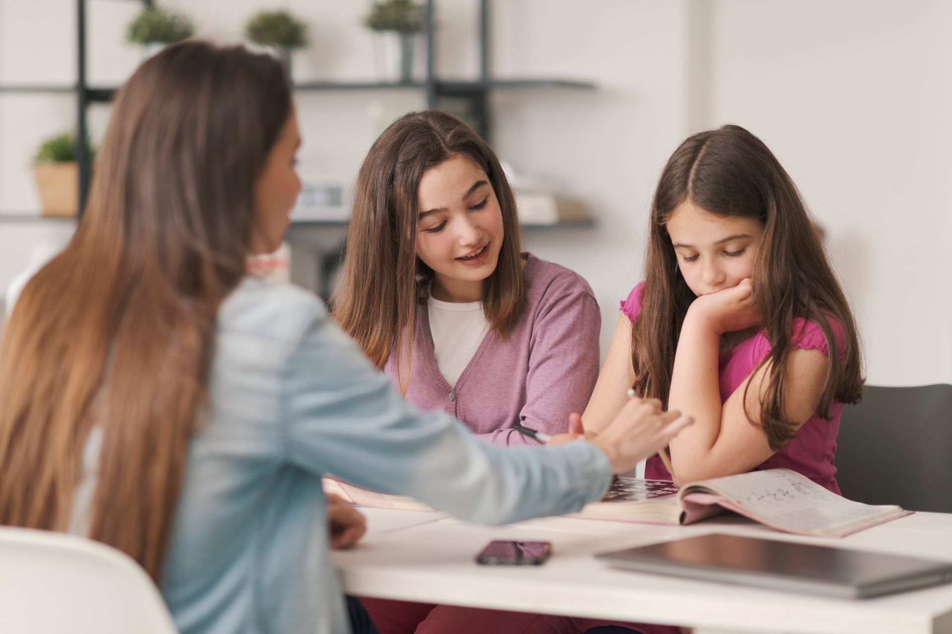 Three people at a table; one points, two look. One child appears thoughtful with a book.