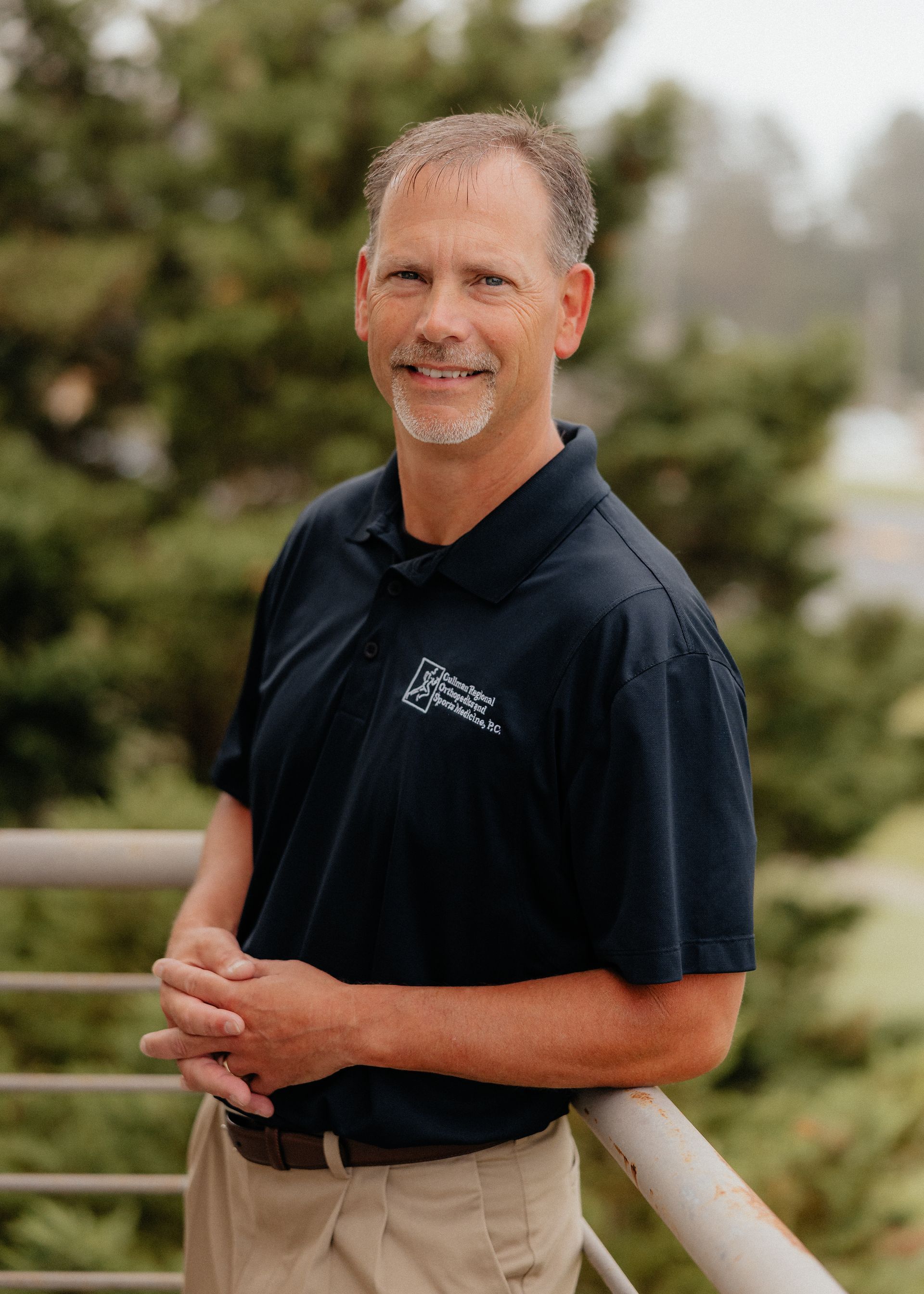Tommy Mullican, PA-C in a blue shirt is leaning against a brick wall with his arms crossed.