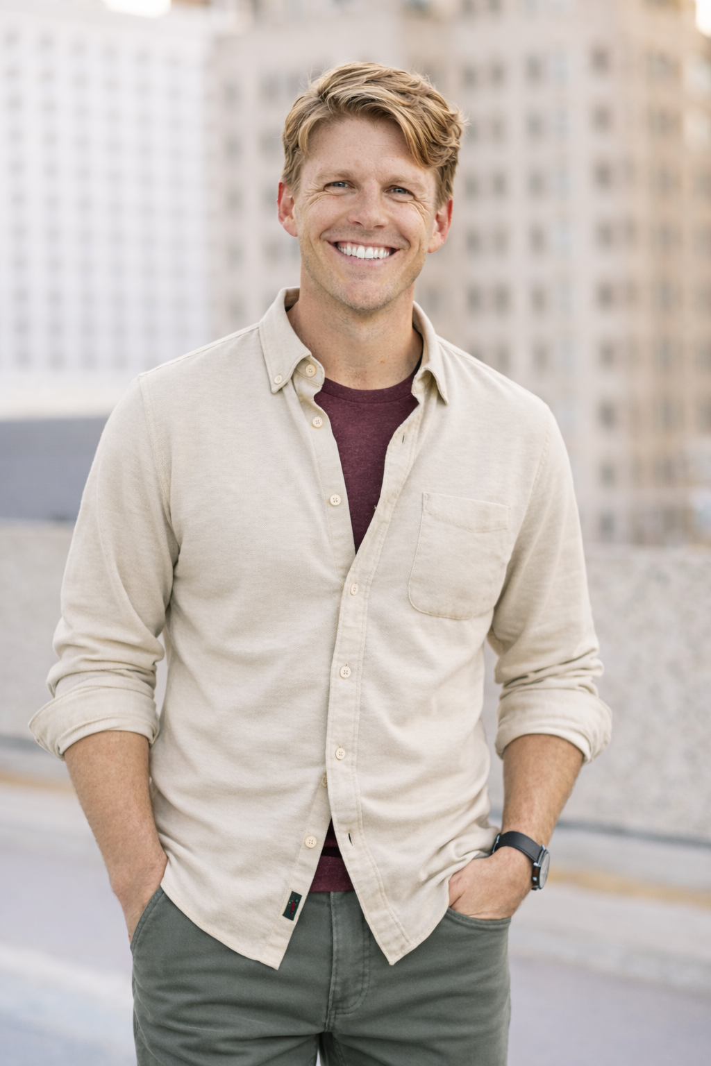A smiling person wearing a beige button-down shirt over a maroon t-shirt and green pants, standing outdoors.