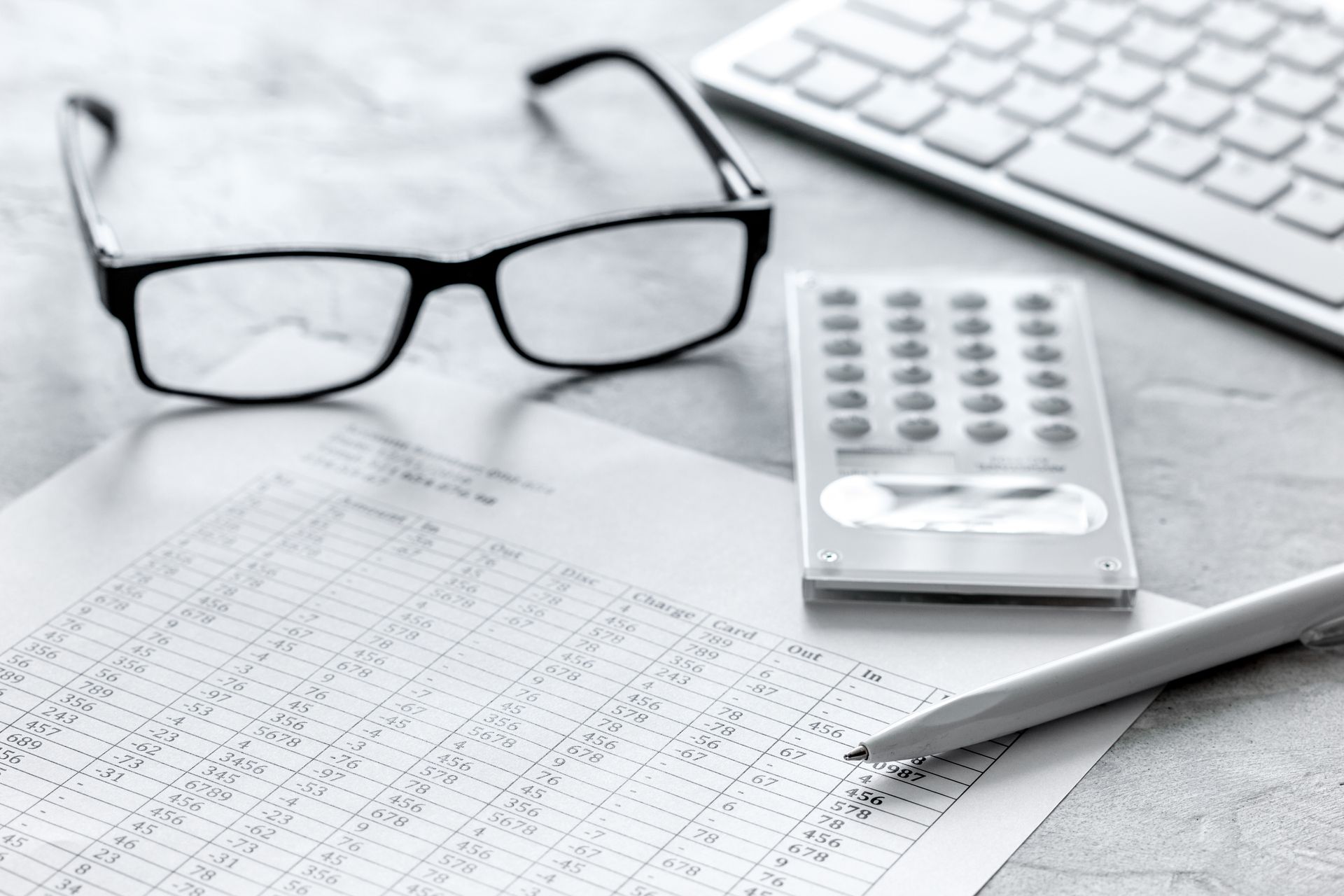 A pair of glasses , a calculator , a pen and a keyboard are on a table.