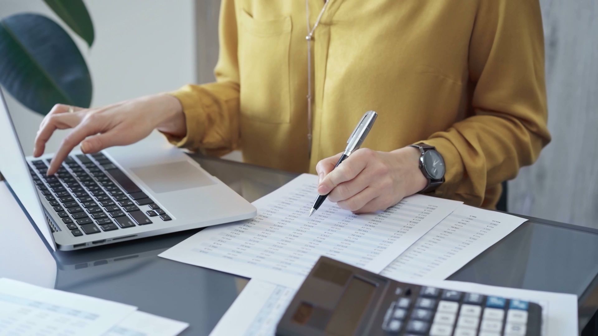 A woman is sitting at a desk using a laptop computer and writing on a piece of paper.