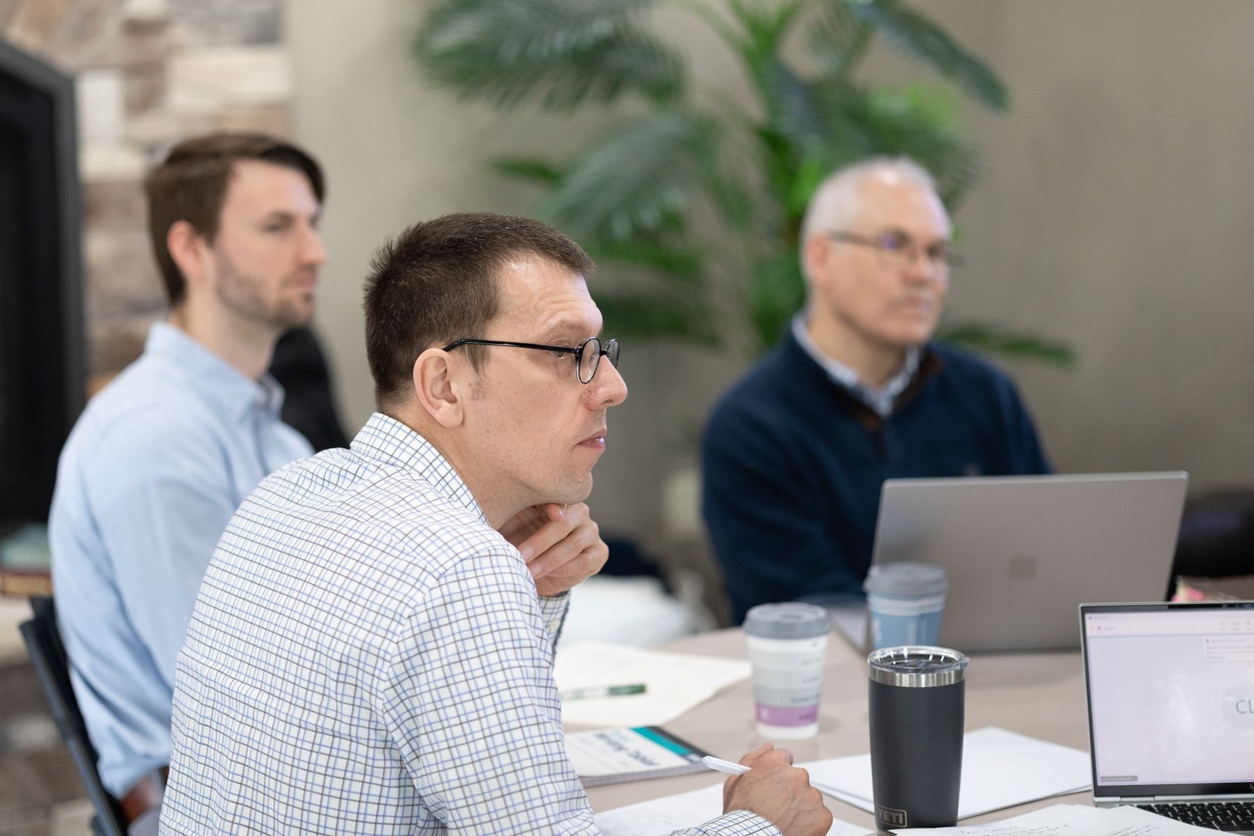 Three men in a meeting, focused on a table with papers, laptops, and coffee.