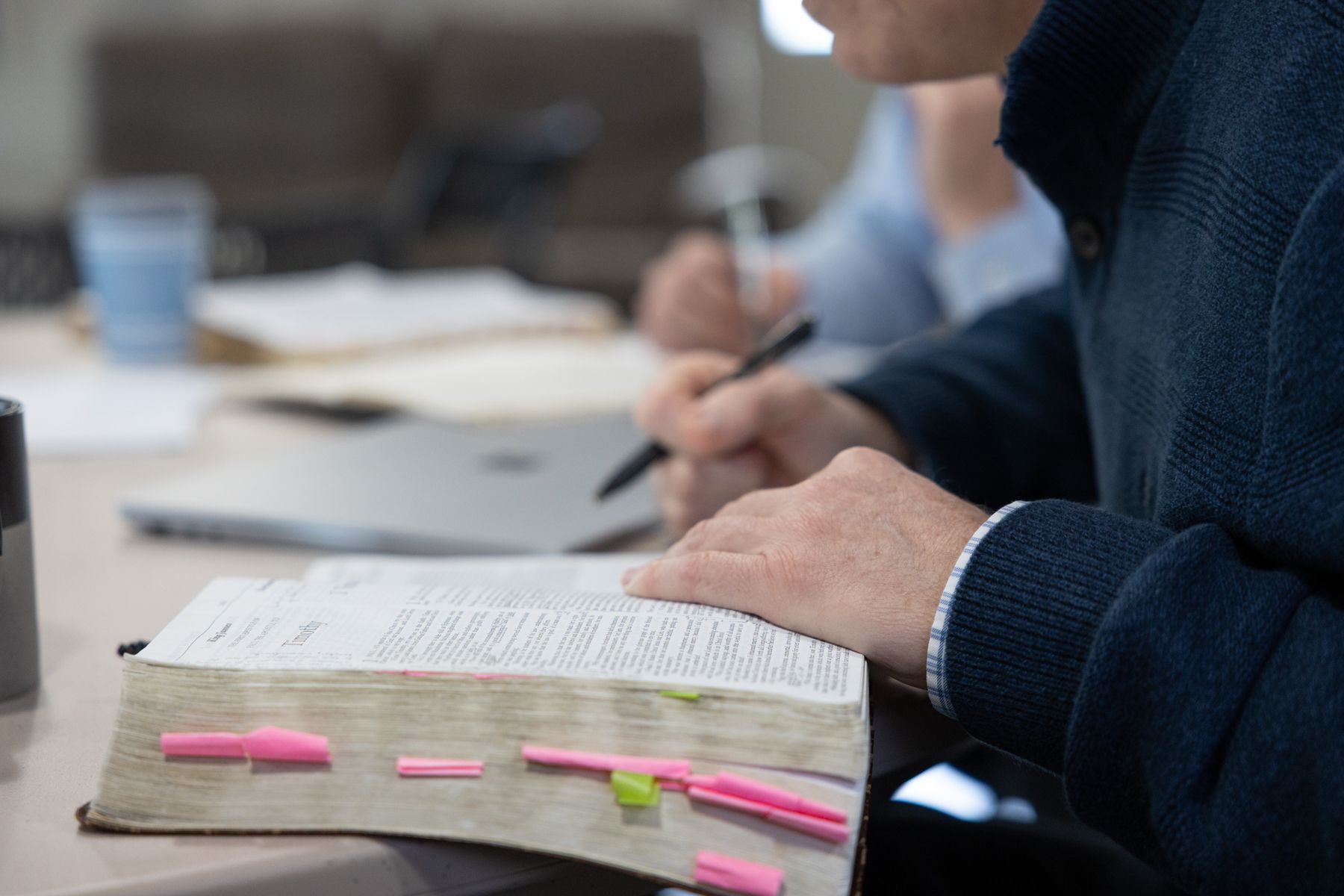A person with a pen writing in a book with highlighted passages, at a table with others.