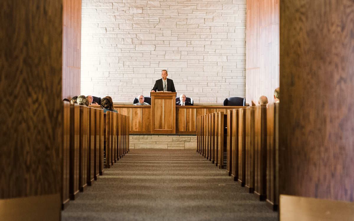 A man stands at a wooden podium in a church, speaking to seated figures. Rows of pews lead toward the speaker.