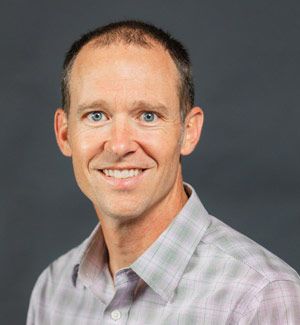 Smiling man in a light plaid shirt, blue eyes, and short brown hair, posing against a gray backdrop.