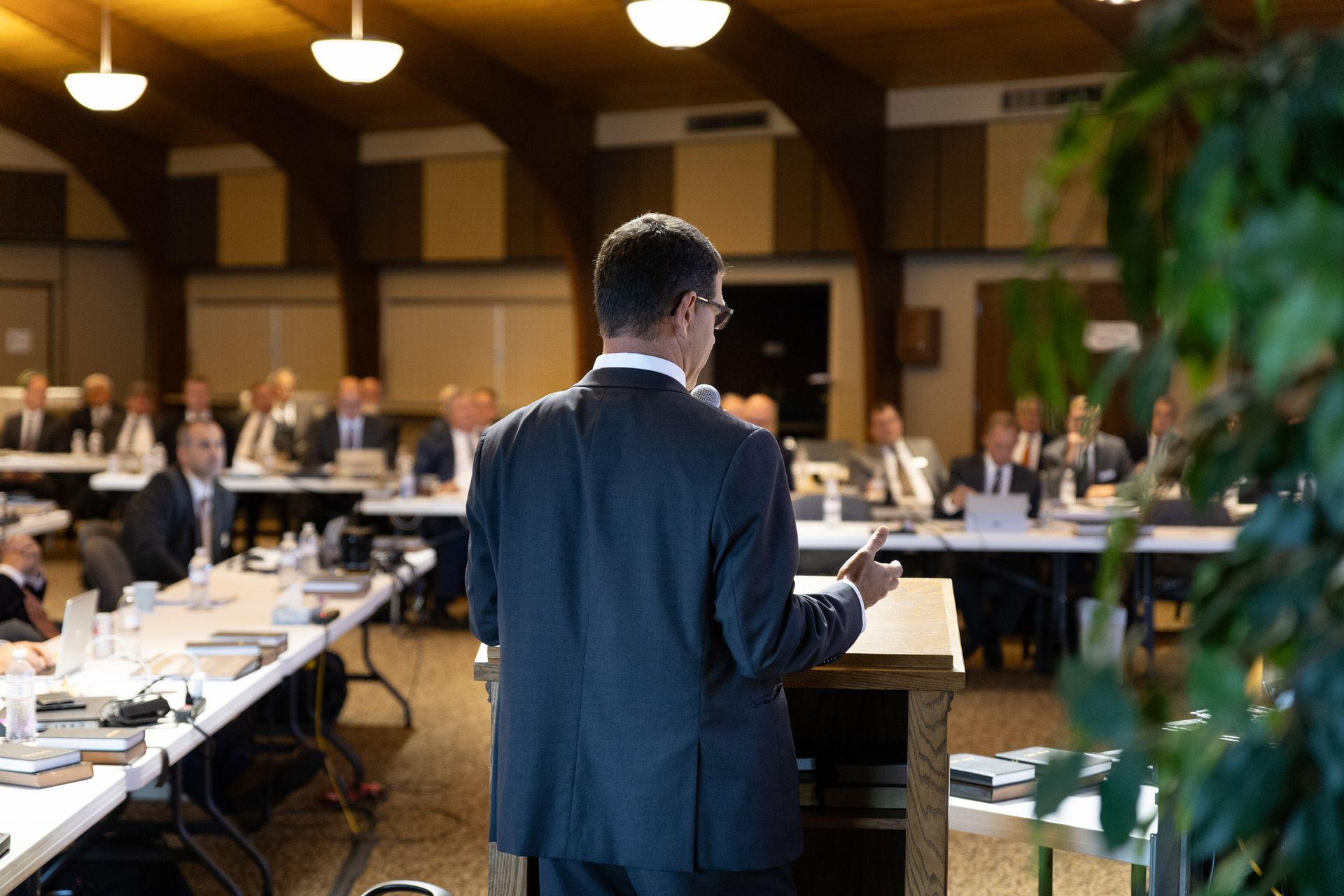 A man in a suit speaks from a podium at a conference. Attendees sit at tables in a large room with wood paneling.