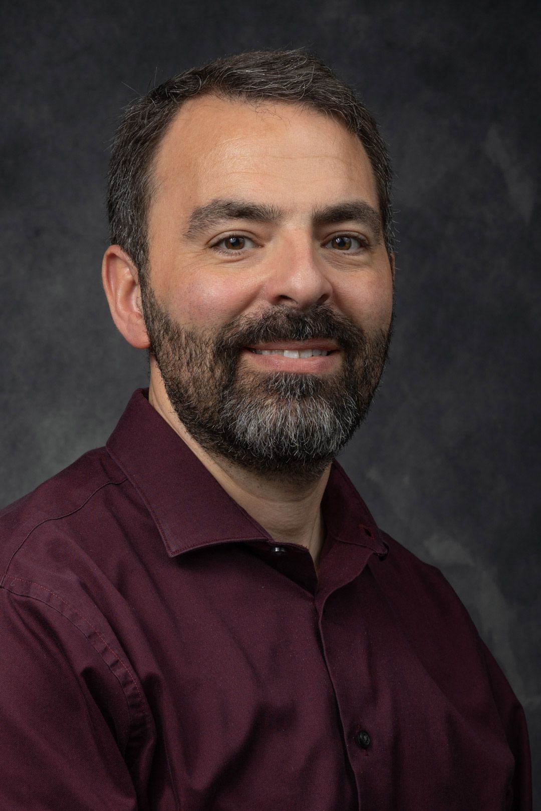 Man with a dark beard and burgundy shirt smiles. He is in a studio setting with a dark background.