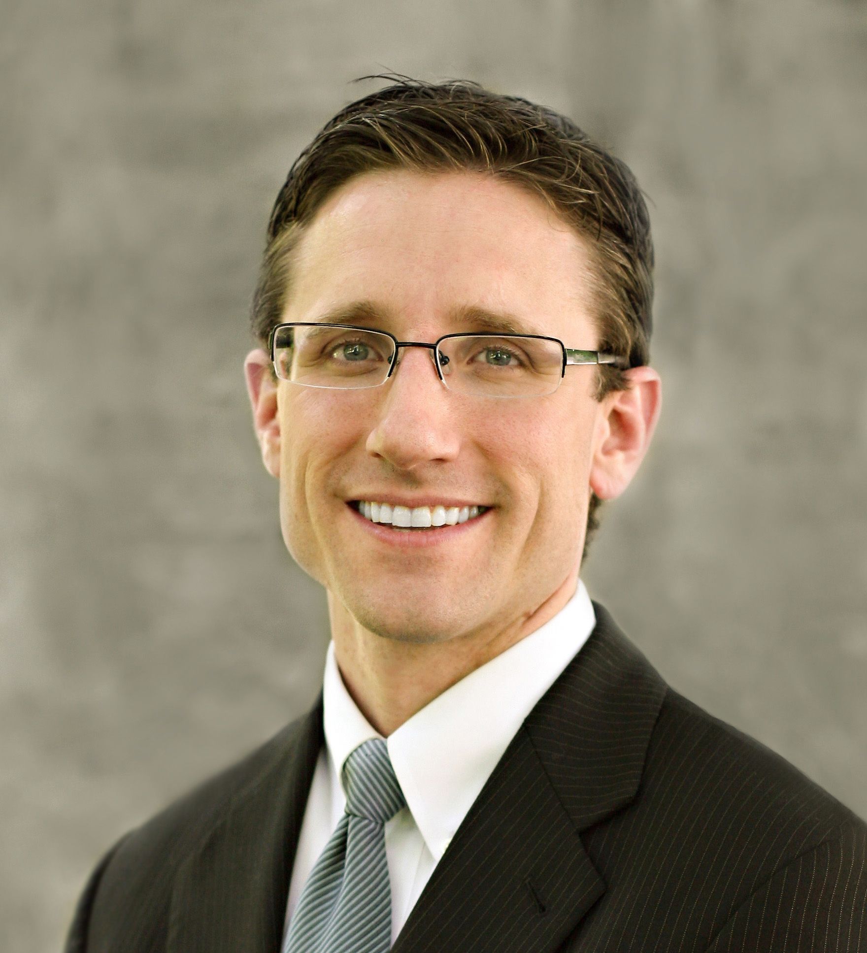 Man in glasses, wearing a suit and tie, smiling broadly against a gray background.