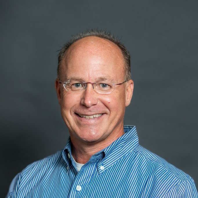 Bald man in glasses, smiling, wearing a blue button-down shirt, against a gray background.