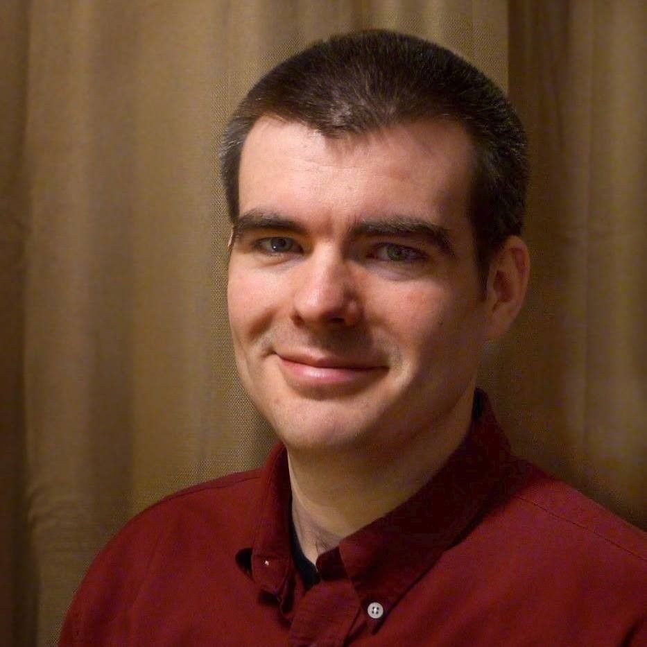 Man with fair skin and short dark hair smiling, wearing a maroon button-up shirt, against a brown curtain backdrop.