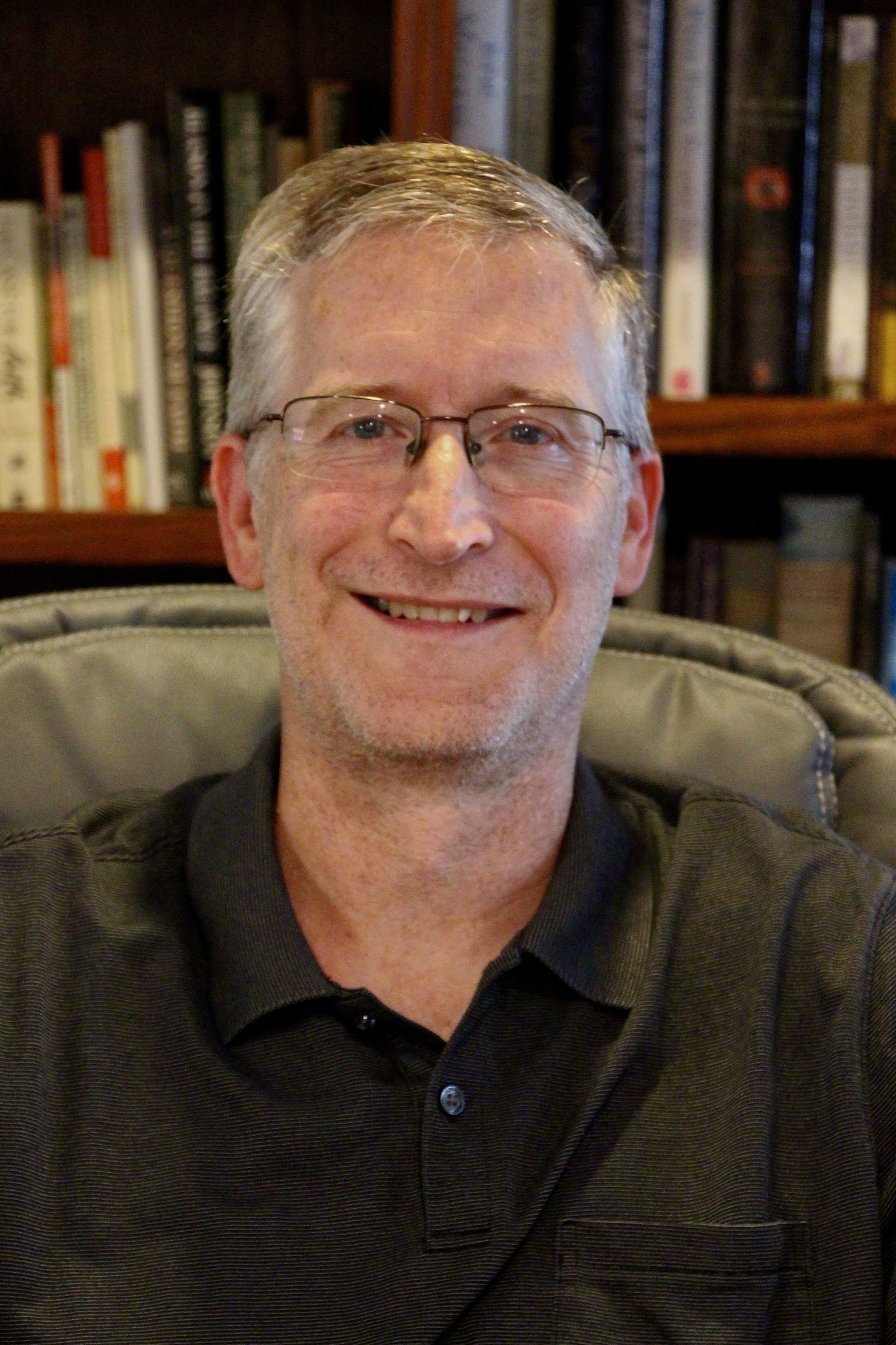 Man with glasses smiles, seated in a chair against a bookshelf backdrop. 