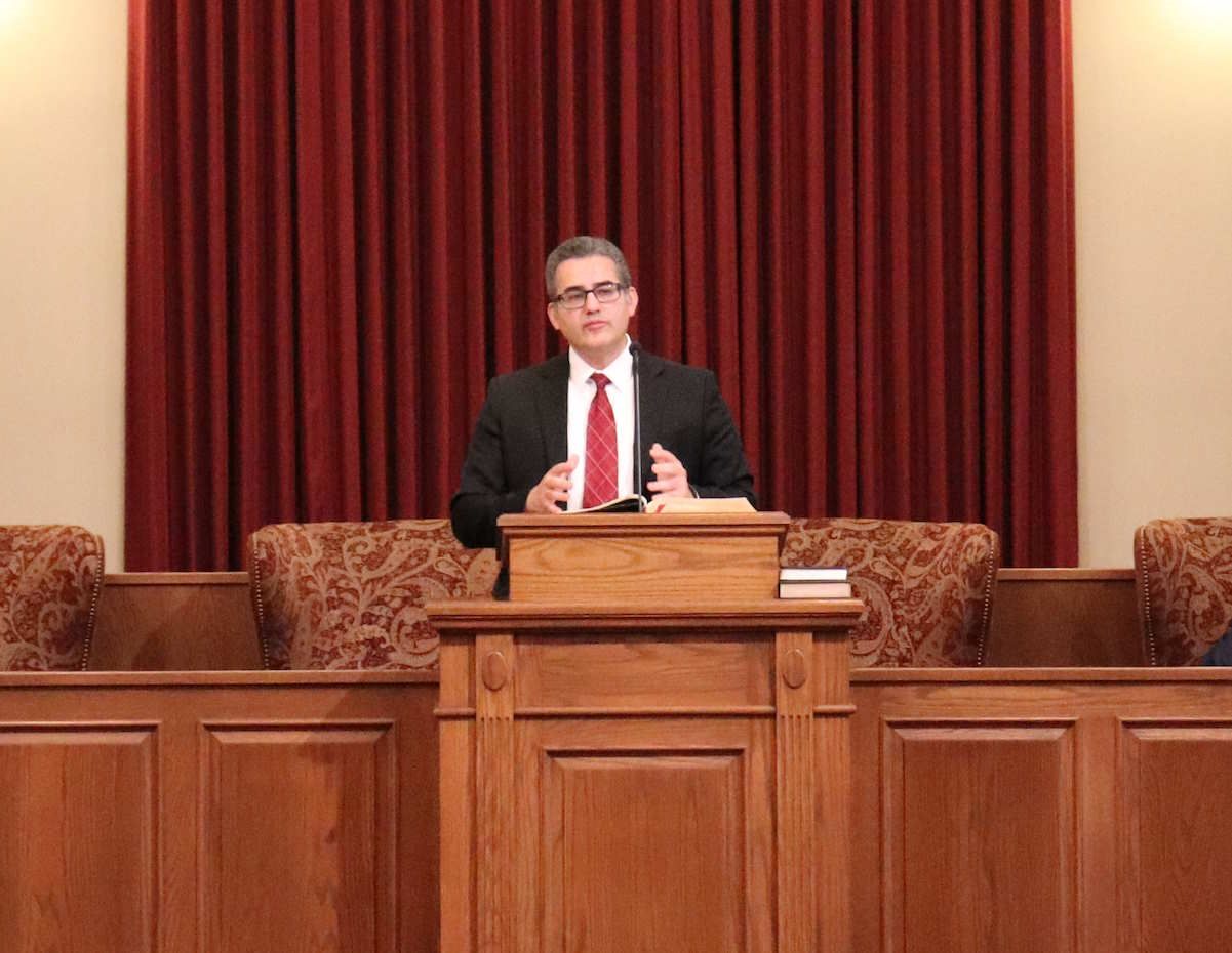 A man in a suit stands at a wooden pulpit in front of a red curtain, speaking with his hands. He is in a church.