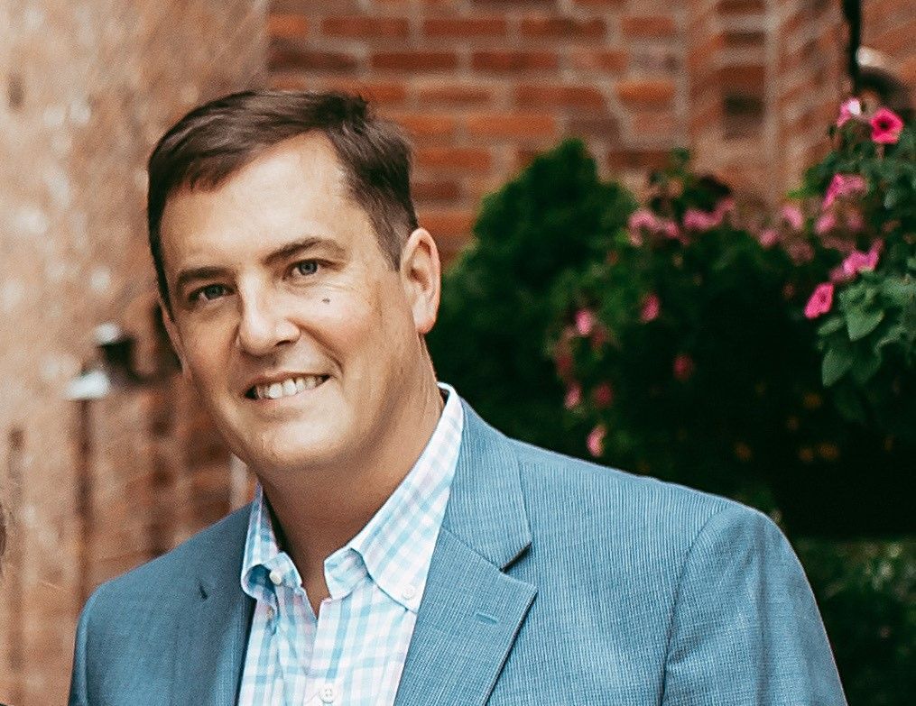 Man in a blue blazer and plaid shirt smiles. He is standing outside near a brick wall and flowers.