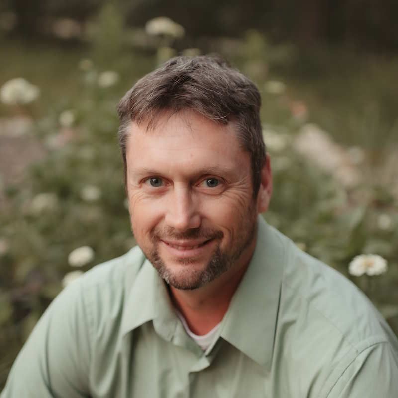 Man in green shirt smiles outdoors, green and white blurred background.