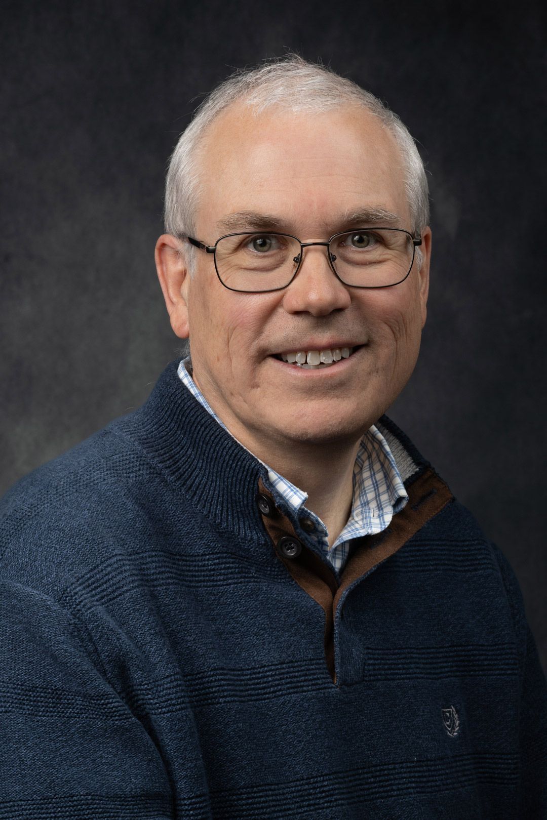 Man with glasses smiling, wearing a blue sweater, against a dark gray background.