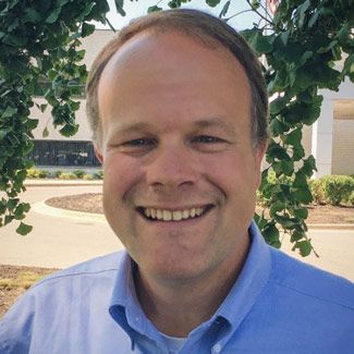 Smiling man in a blue shirt, standing outdoors with green foliage and a building in the background.