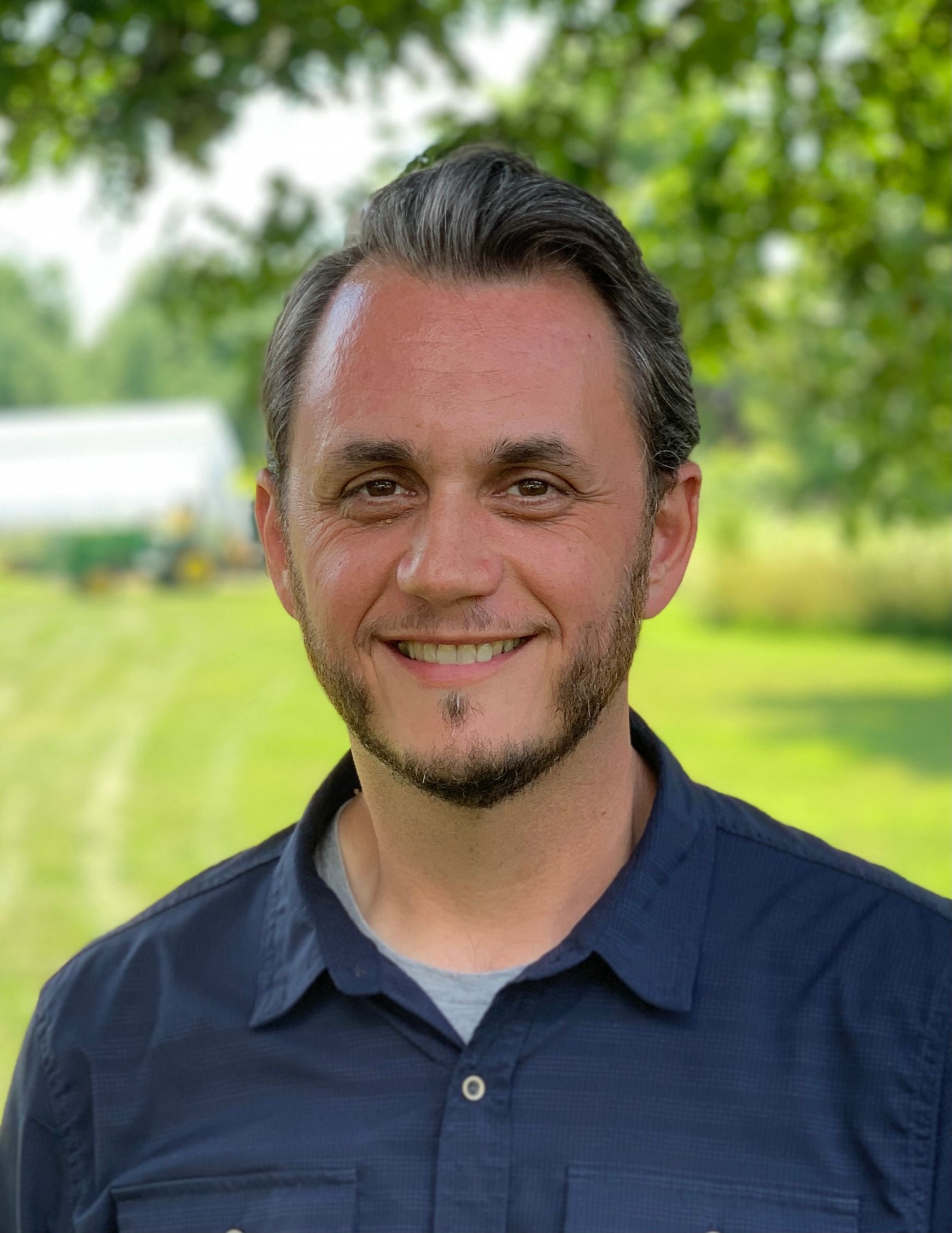 Smiling man with dark hair and beard in a blue shirt, set outdoors. A greenhouse is visible in the background.