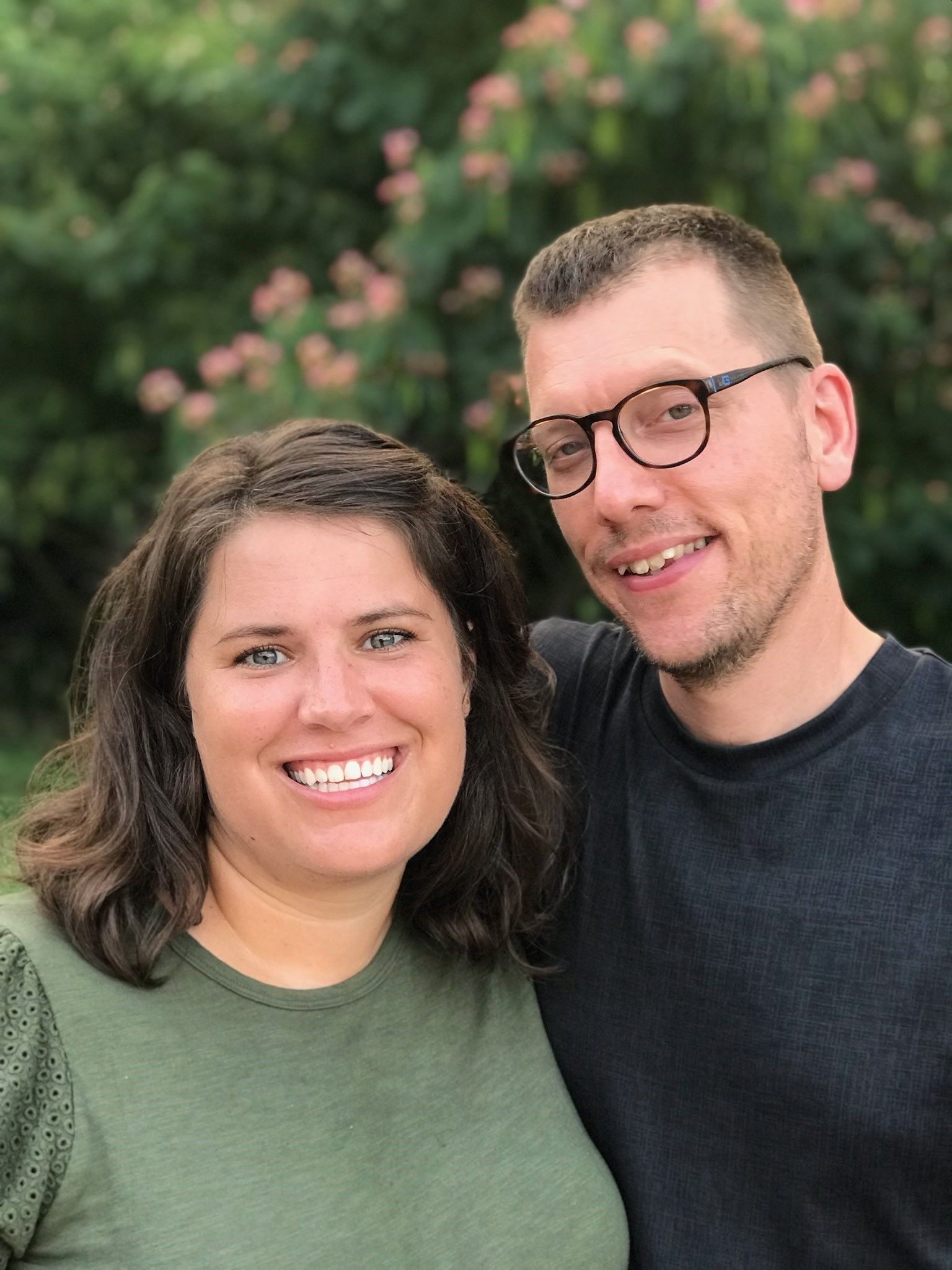 A man and woman smiling together outdoors. The man wears glasses, a suit and tie, while the woman wears a black shirt.