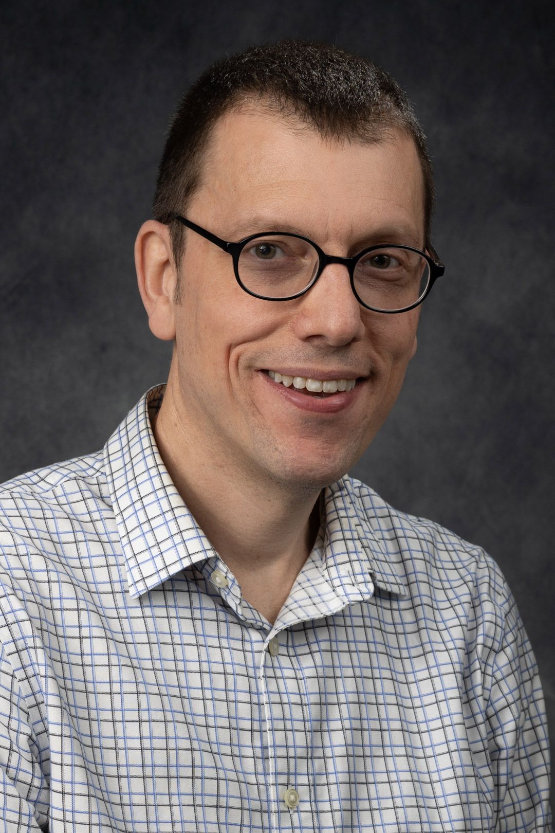 Man with glasses smiles at the camera, wearing a button-down shirt against a dark background.