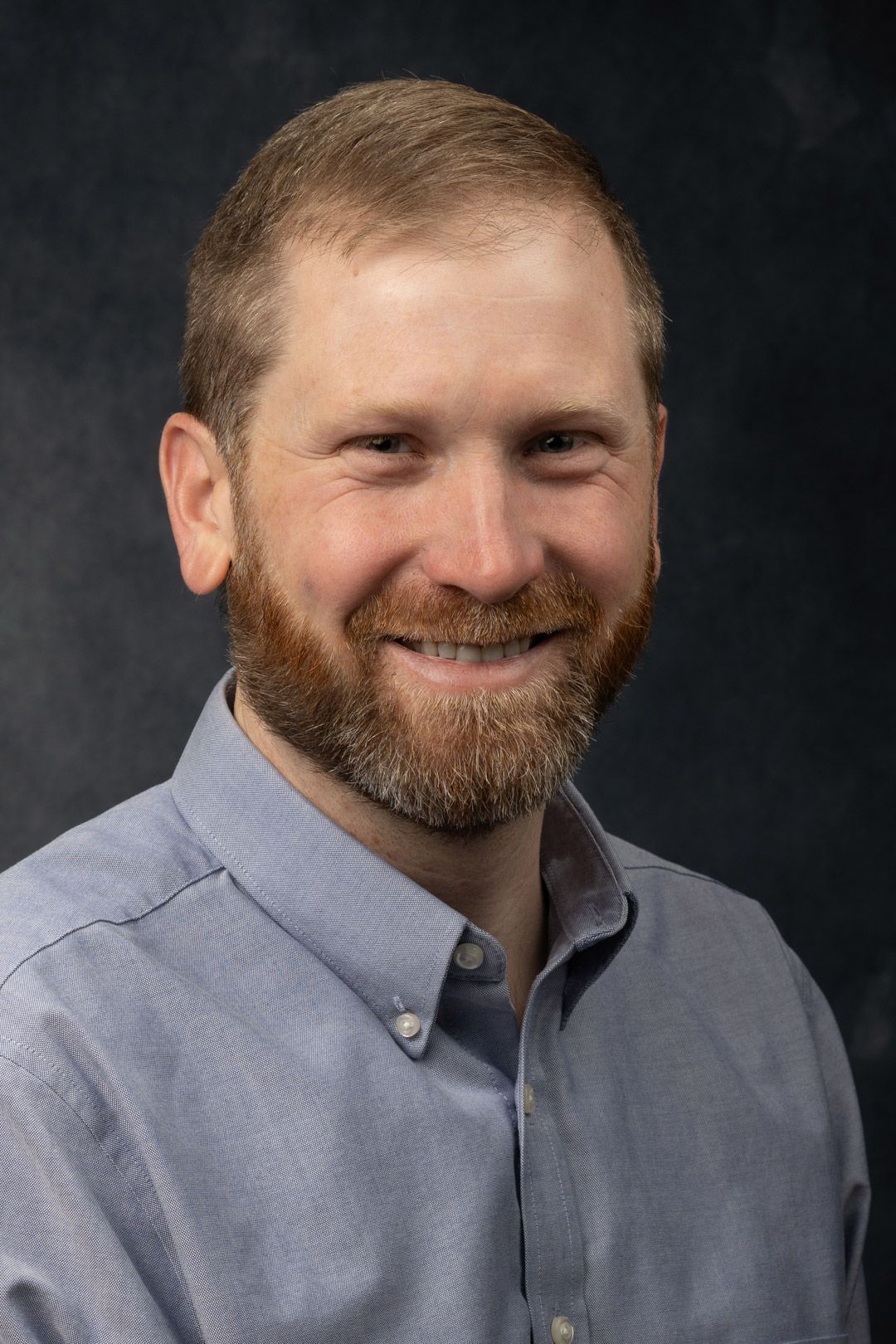 Man with reddish-brown beard smiling, wearing a light blue button-down shirt, with a dark background.