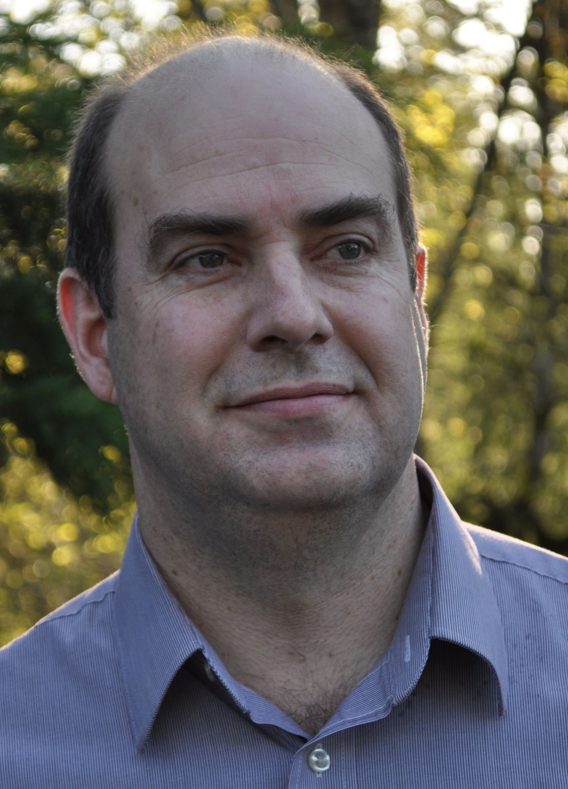 Bald man with fair skin, wearing a blue button-down shirt, smiles gently outdoors with blurred foliage in the background.