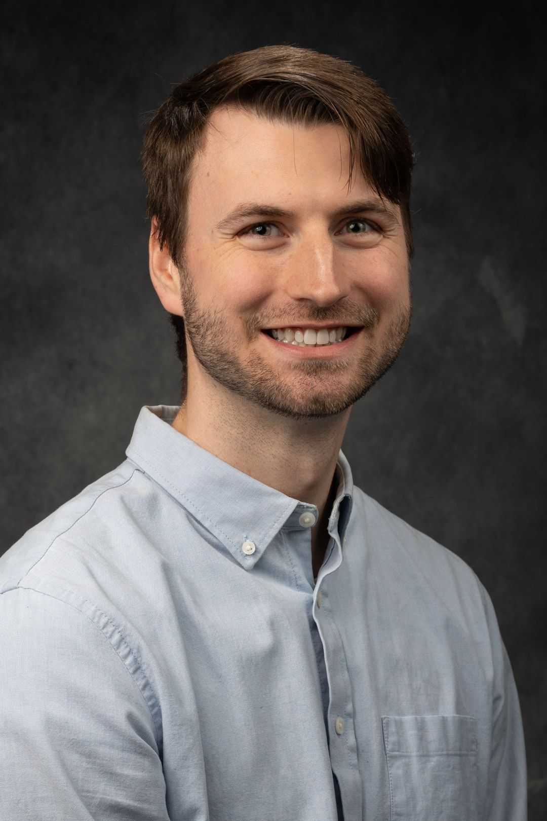 Smiling man with light brown hair and stubble, wearing a light blue button-down shirt against a dark gray backdrop.