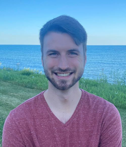 Smiling man with a light beard in a red shirt, standing in front of a grassy area, with the ocean and sky in the background.