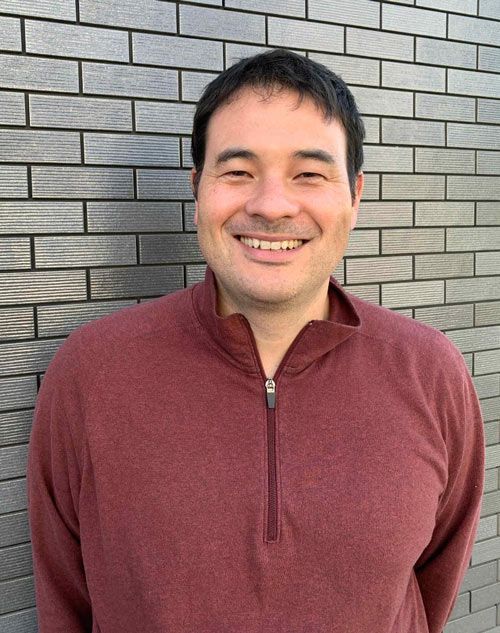 Smiling man in a maroon quarter-zip shirt stands in front of a gray brick wall.