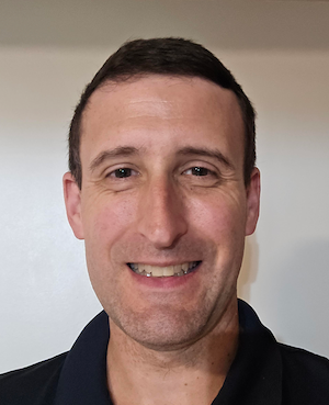 A man with short brown hair smiles at the camera. He is wearing a dark shirt against a white background.