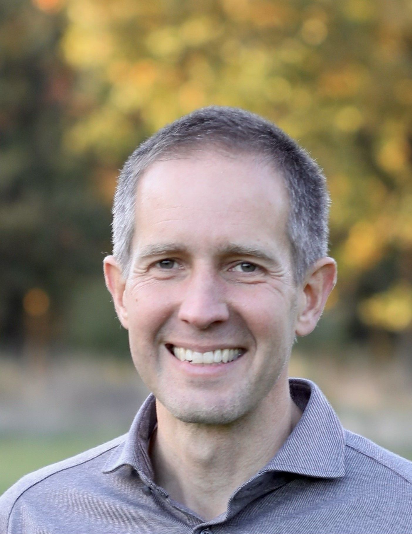 Smiling man with short gray hair outdoors, wearing a gray shirt.