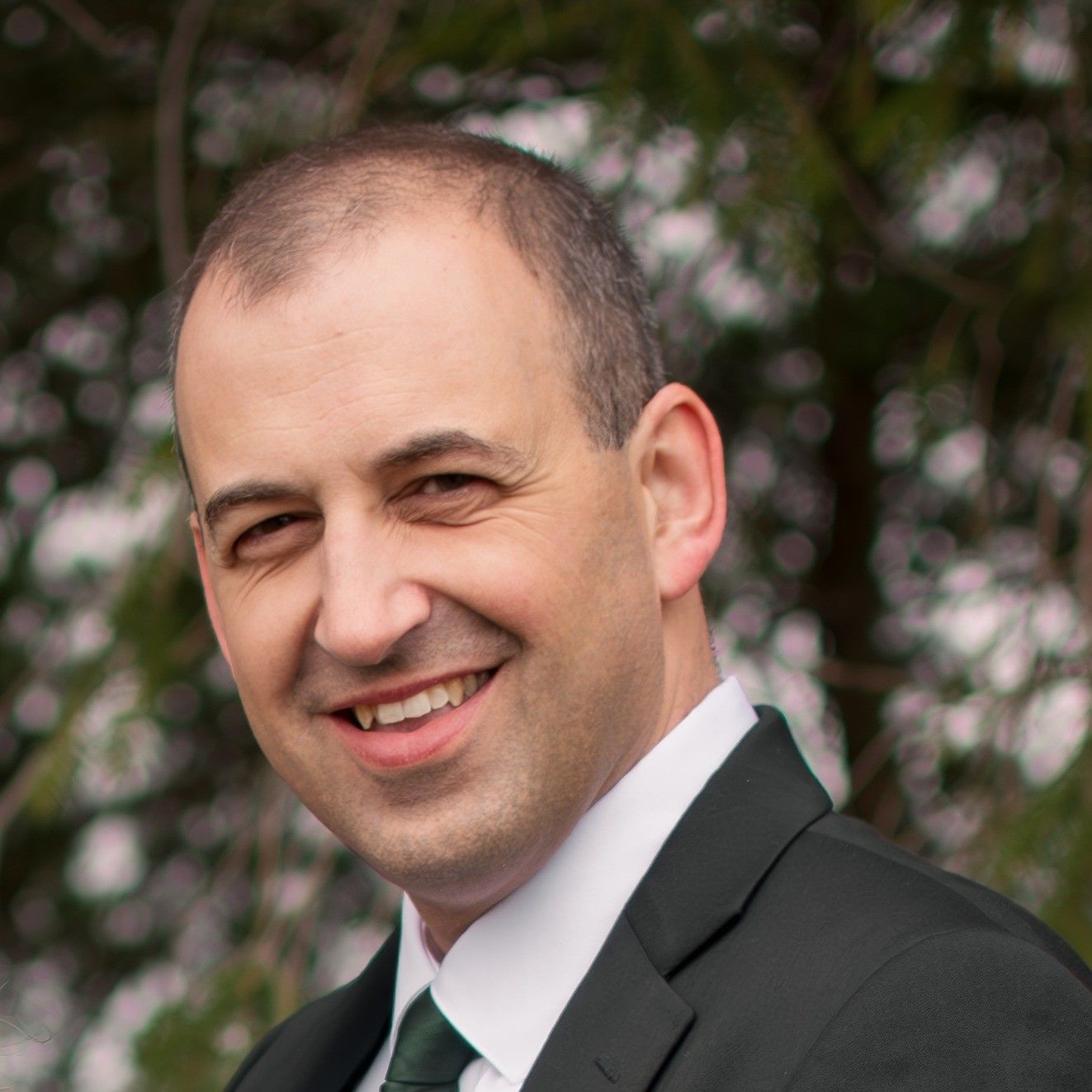 Man with short brown hair, smiling, wearing a black suit with a green tie, outdoors.