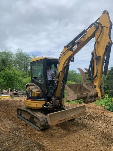 A man is driving a small yellow excavator in a dirt field.