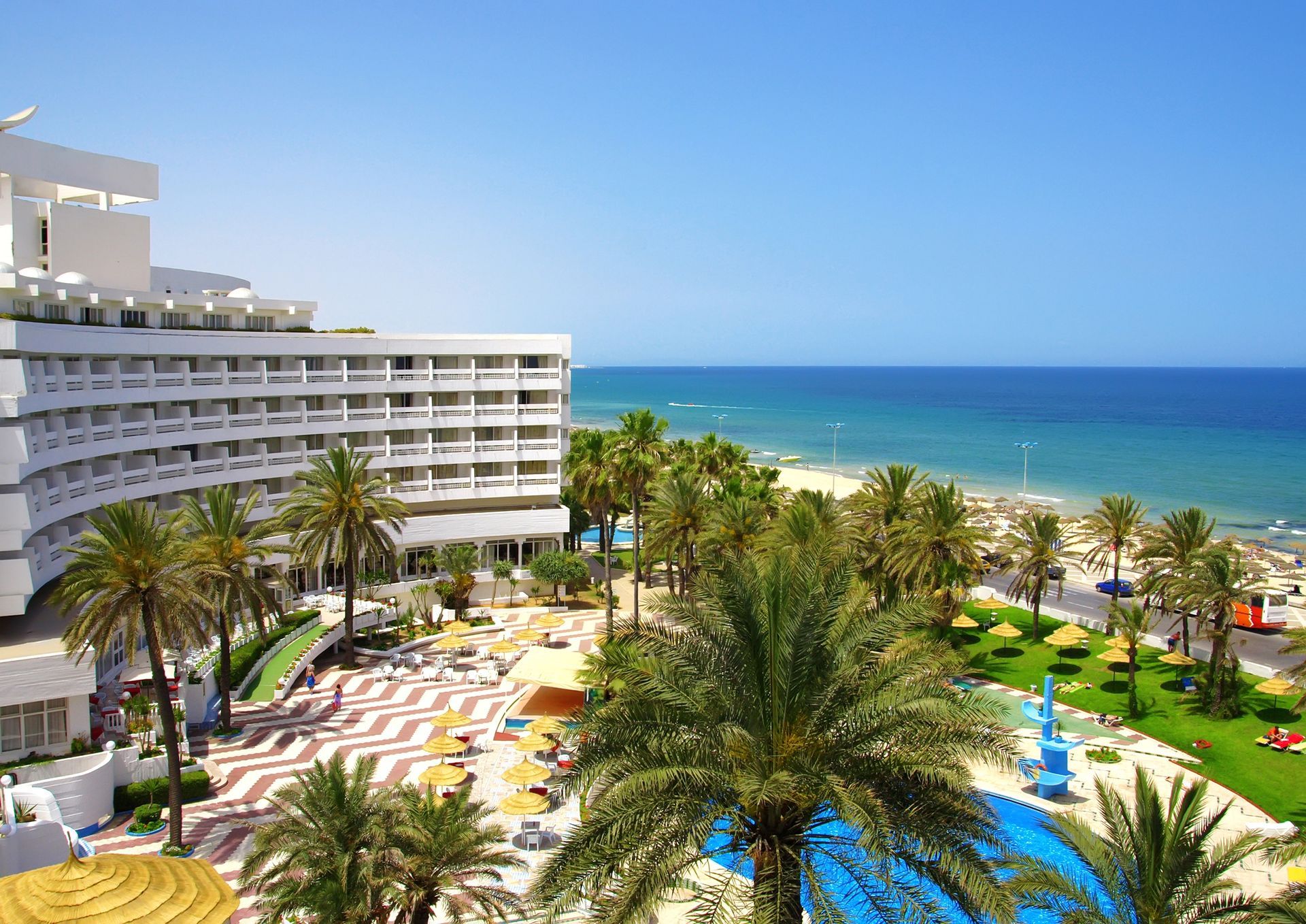Resort hotelero de color blanco con vistas a la playa y al océano con palmeras bajo un cielo azul.