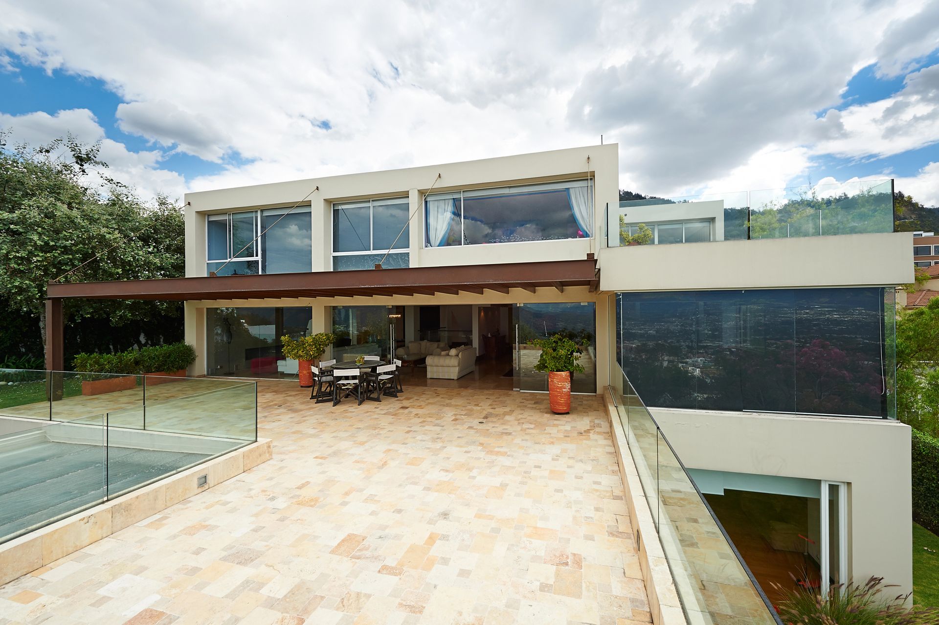 Exterior de casa moderna con amplios ventanales, patio y vista parcial a una cancha de tenis, bajo un cielo nublado.
